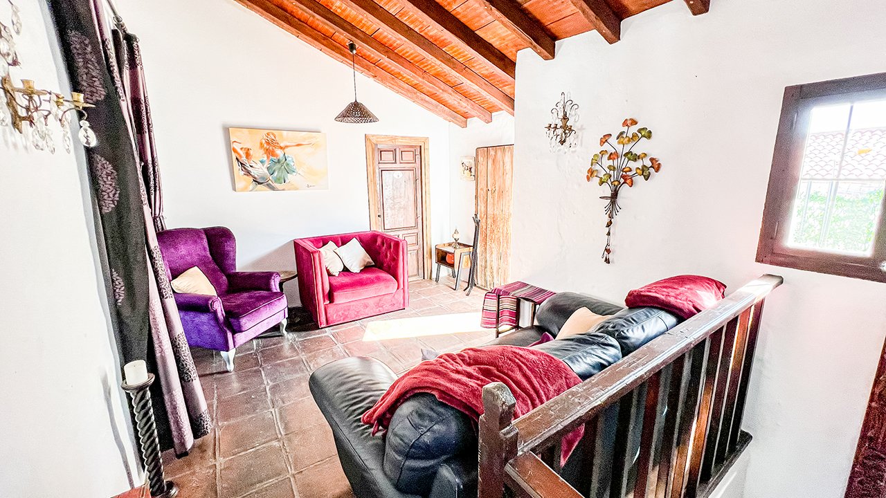 Living room with white walls, wooden ceiling beams, and a tiled floor. Contains a purple armchair, a red armchair, and a black leather couch with red blankets. There are framed art, wall-mounted candleholders, a decorative wall hanging, and a window 