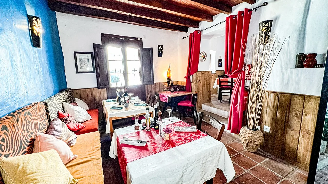 A cozy dining room with a long dining table covered with a white and red tablecloth, set with plates, glasses, and condiments. The room features a blue and white wall with wooden accents, red curtains, and a large window letting in natural light. The