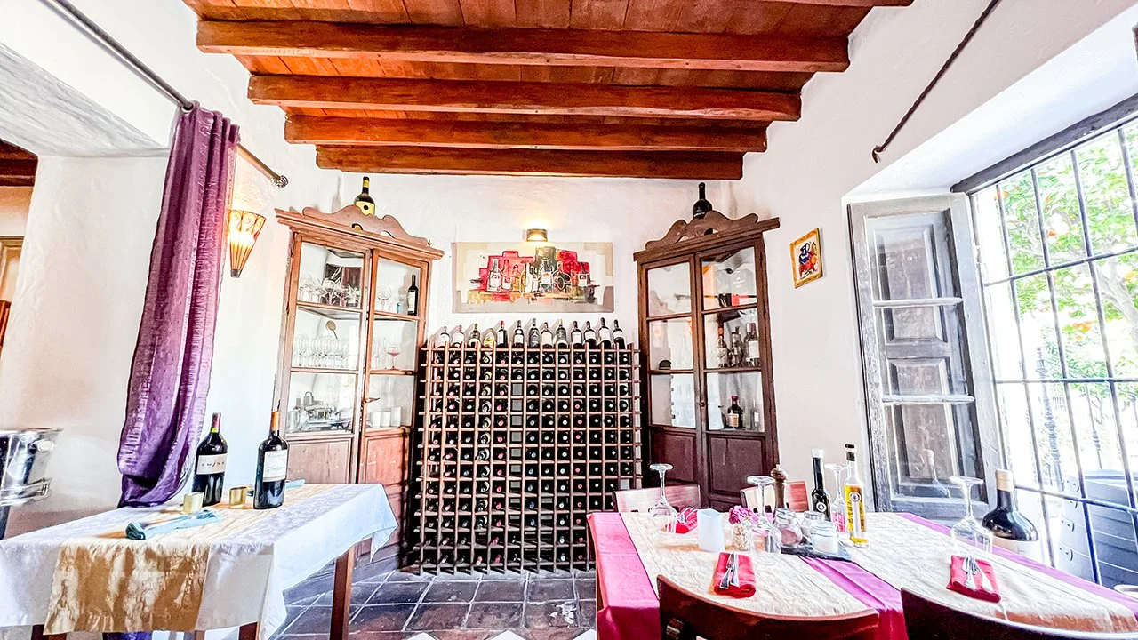 Interior of a dining room with wooden ceiling beams, a large window with metal bars showing greenery outside, and a central wine rack with an assortment of wine bottles. There are cabinets with glass doors, decorative wine bottles on top, and a dinin