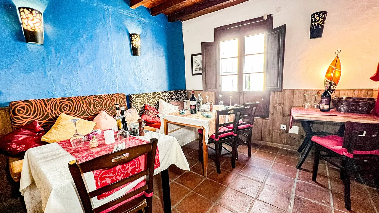 Dining area with colorful cushions, bottles on the table, and a window letting in bright sunlight in a rustic-style room
