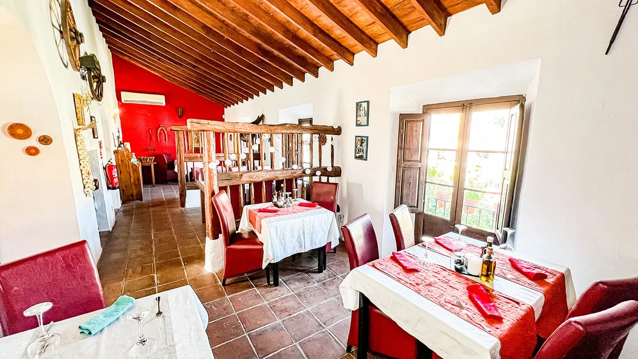 Interior of a restaurant with white walls, red accent wall, wooden ceiling beams, and large window with shutters. There are tables with white tablecloths, red cloth runners, red napkins, and glassware. Red upholstered chairs are around the tables. Th