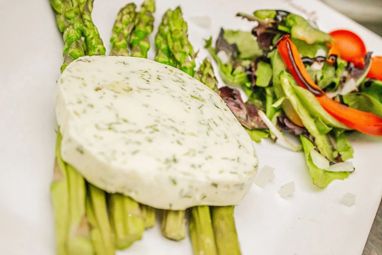 Fresh green asparagus, a wedge of herb-infused cheese, and a mixed green salad with grape tomatoes on a white plate.