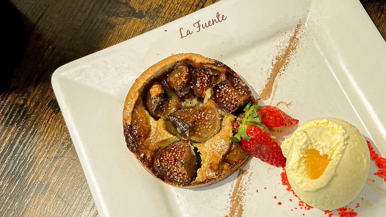 A dessert plate with a fruit tart, garnished with strawberries and a scoop of vanilla ice cream with a fruit topping, on a white square dish with the text "La Fuente".