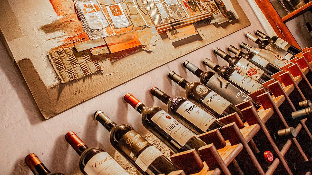 A row of wine bottles displayed on a wooden shelf against a textured wall with an abstract painting hanging above.