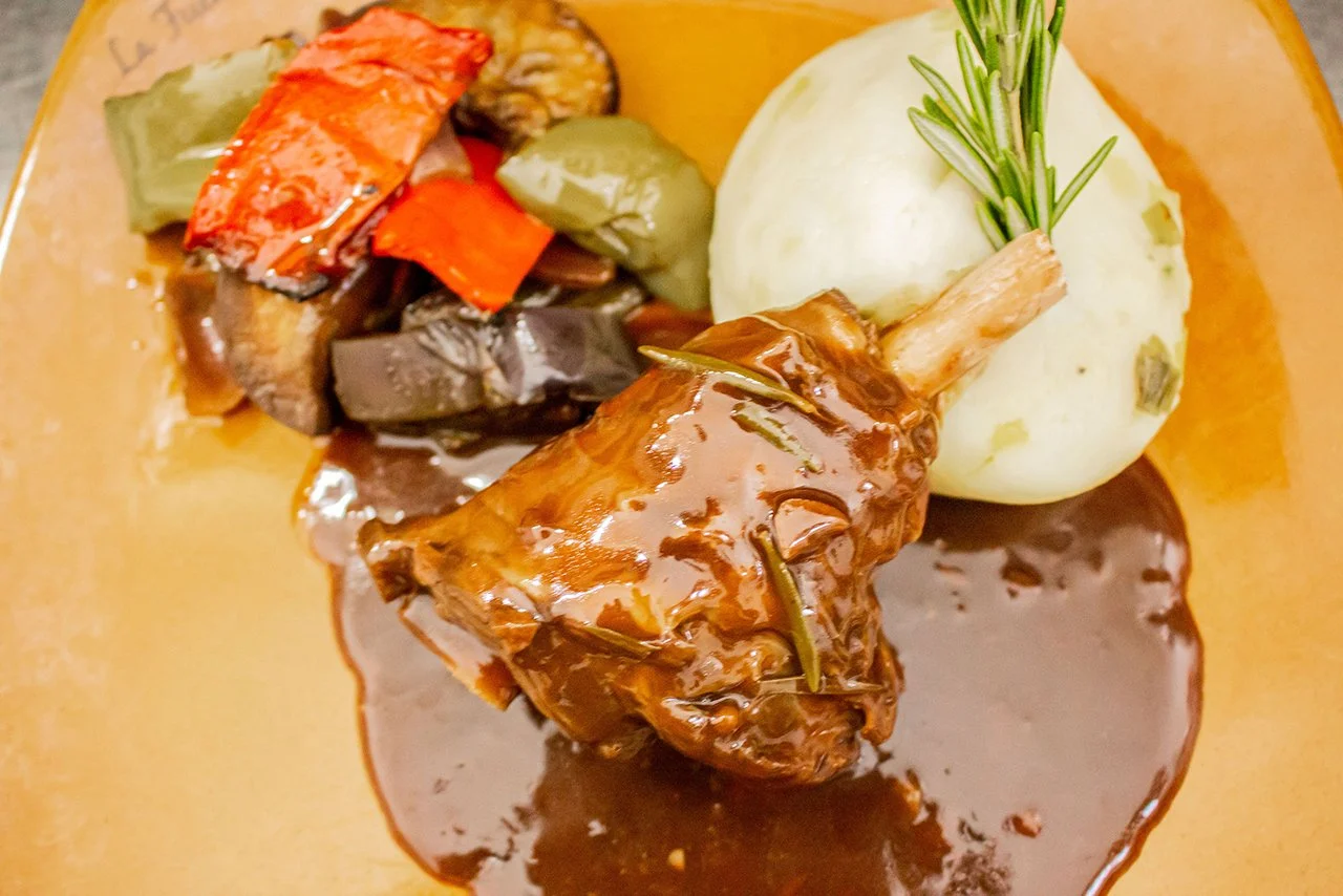 A plate of food featuring a roasted lamb shank with rosemary, mashed potatoes, and a medley of roasted vegetables including eggplant, red bell pepper, and zucchini, all served with a brown gravy.