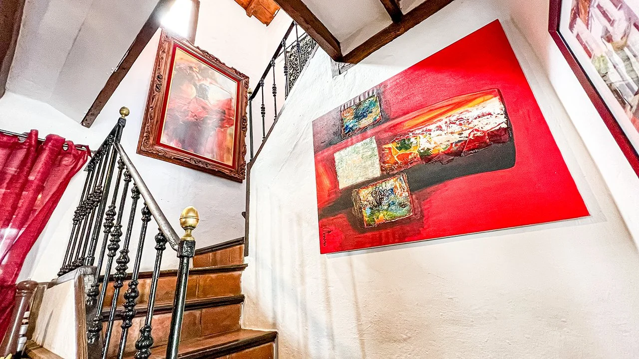 Inside a house, a staircase with black and gold balusters and wooden steps. The wall beside the stairs has two colorful abstract paintings, one large red painting and one framed artwork above the stairs.
