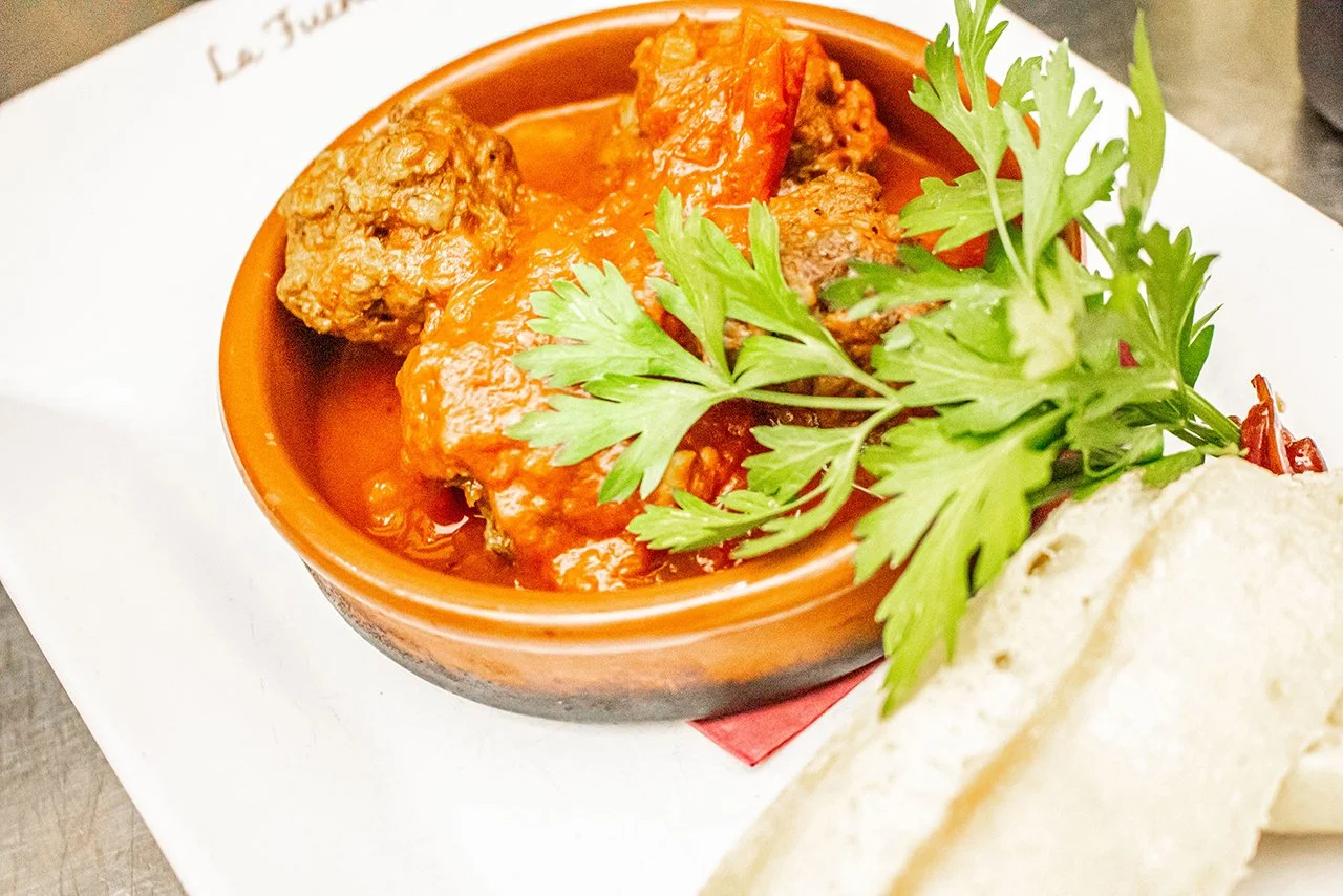 A bowl of meatball stew garnished with parsley, served with bread on a white plate.