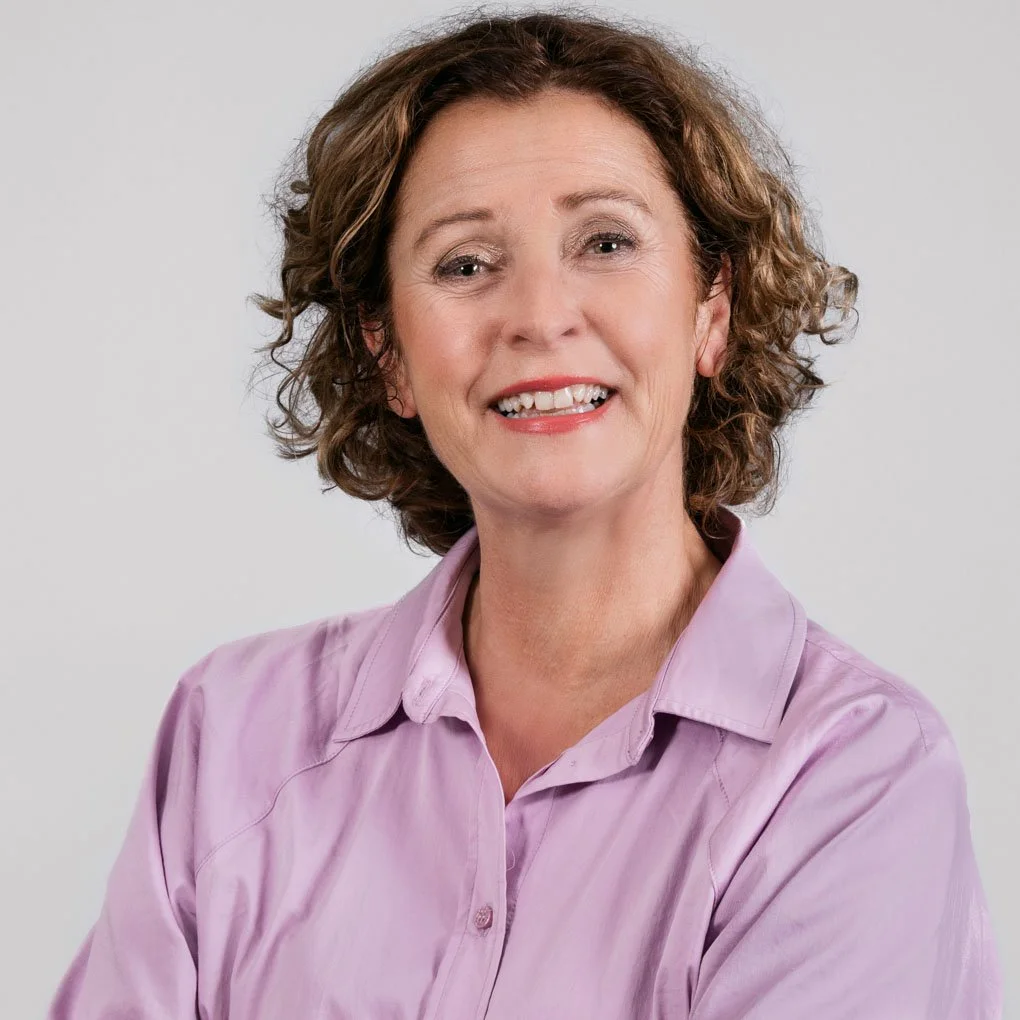 Portret van een vrouw met kort, golvend bruin haar, lachend met zichtbare tanden, gekleed in een lichtpaarse blouse, tegen een lichte grijze achtergrond.