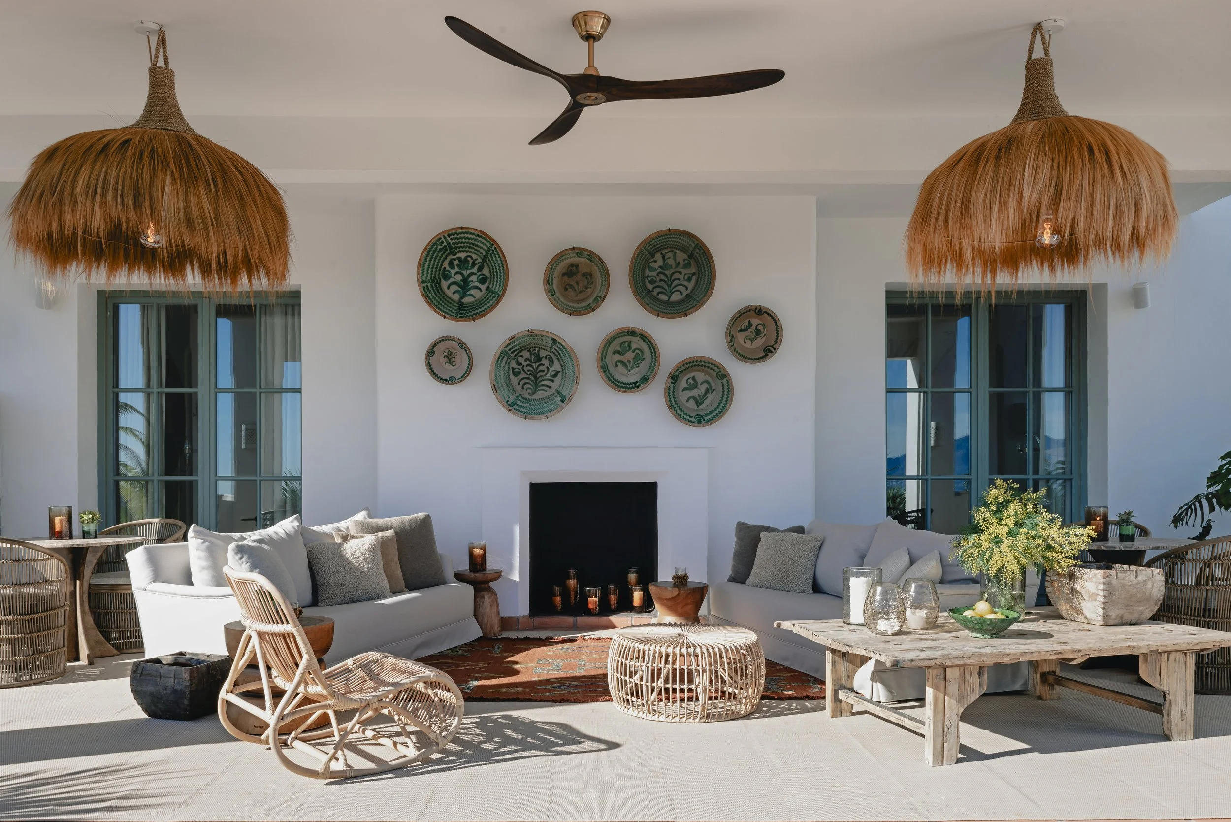A bright, airy outdoor living space with white sofas, wicker chairs, a wooden coffee table, and decorative plates on the wall. Two hanging pendant lights with ruffled shades and a ceiling fan are also visible.