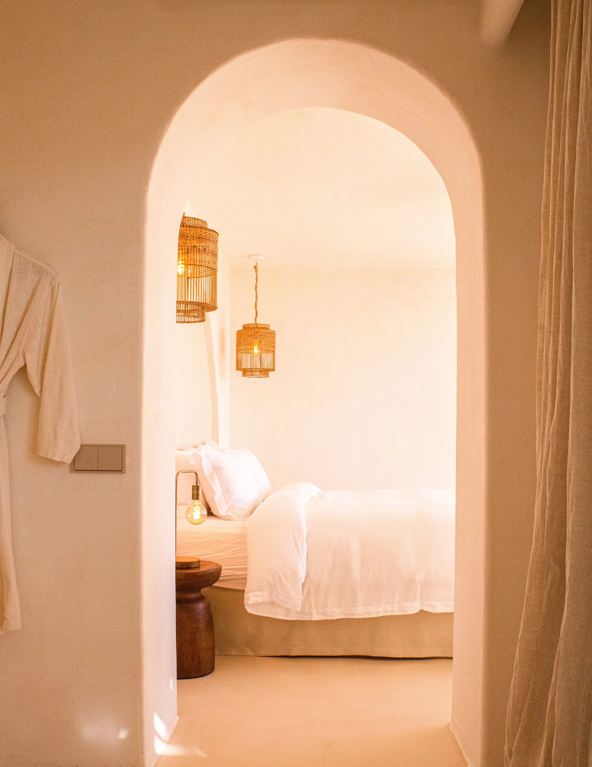 A cozy bedroom with white bed linens, a bedside lamp with an Edison bulb, and woven pendant lighting hanging from the ceiling, seen through an arched doorway with soft lighting.