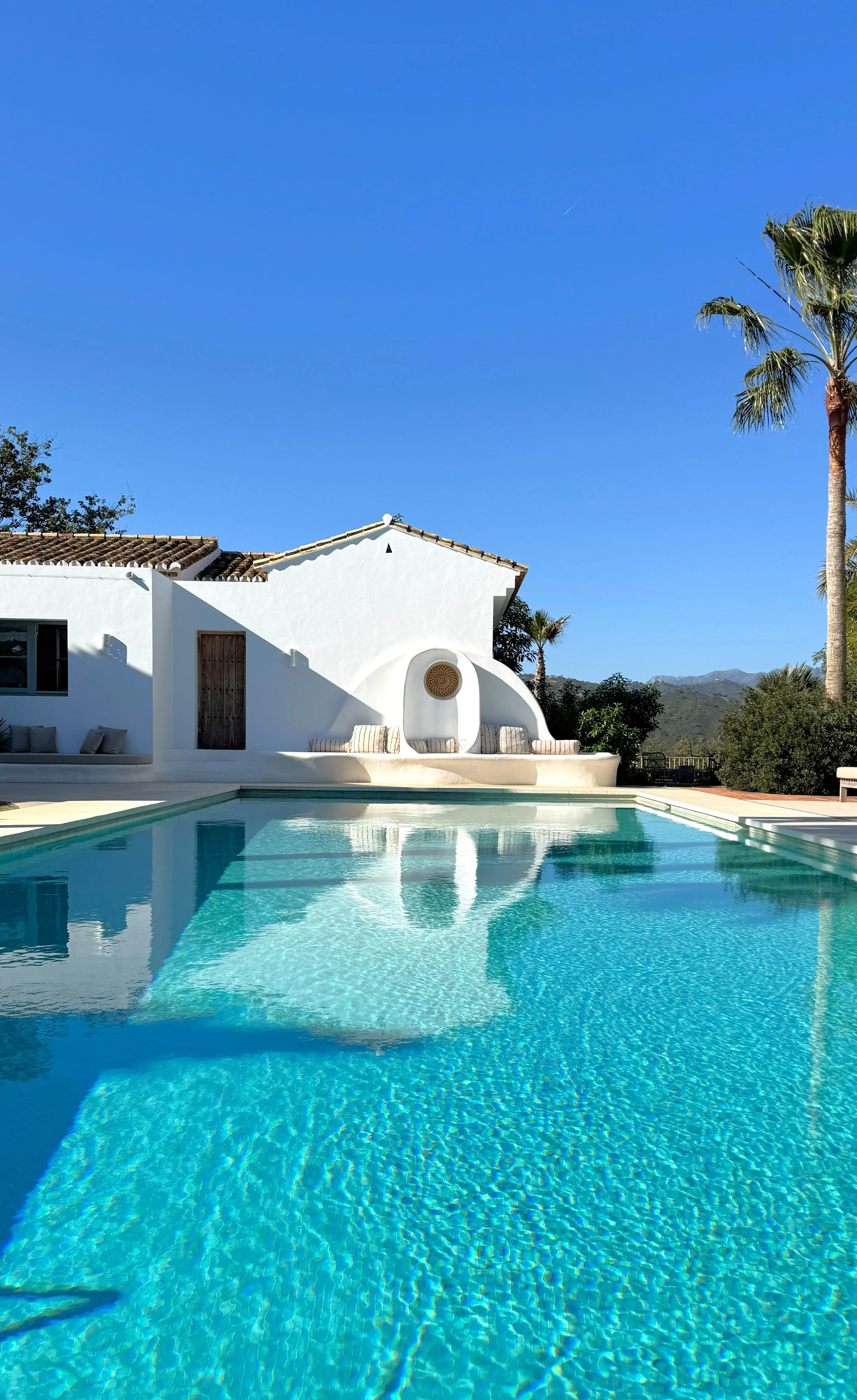 View of a white house with a swimming pool in the foreground, tall palm trees, and mountains in the background under a clear blue sky.
