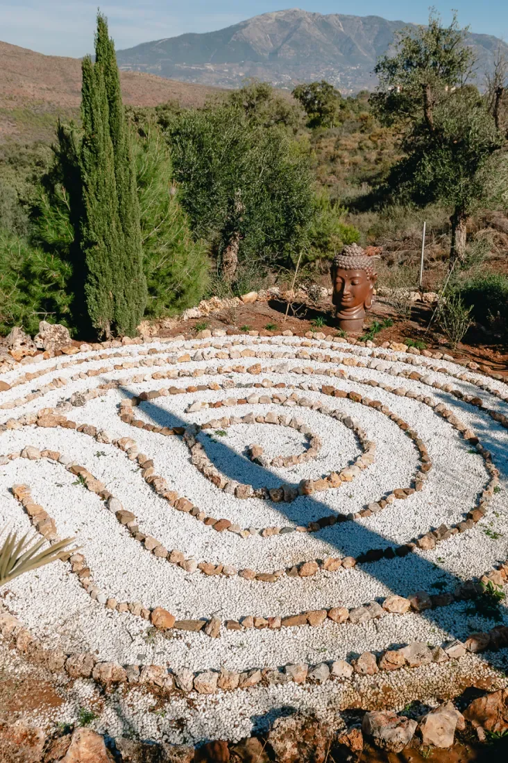 A spiral rock garden with white gravel and rocks, surrounded by green trees and shrubs, with a mountain in the background and a Buddha head statue nearby.