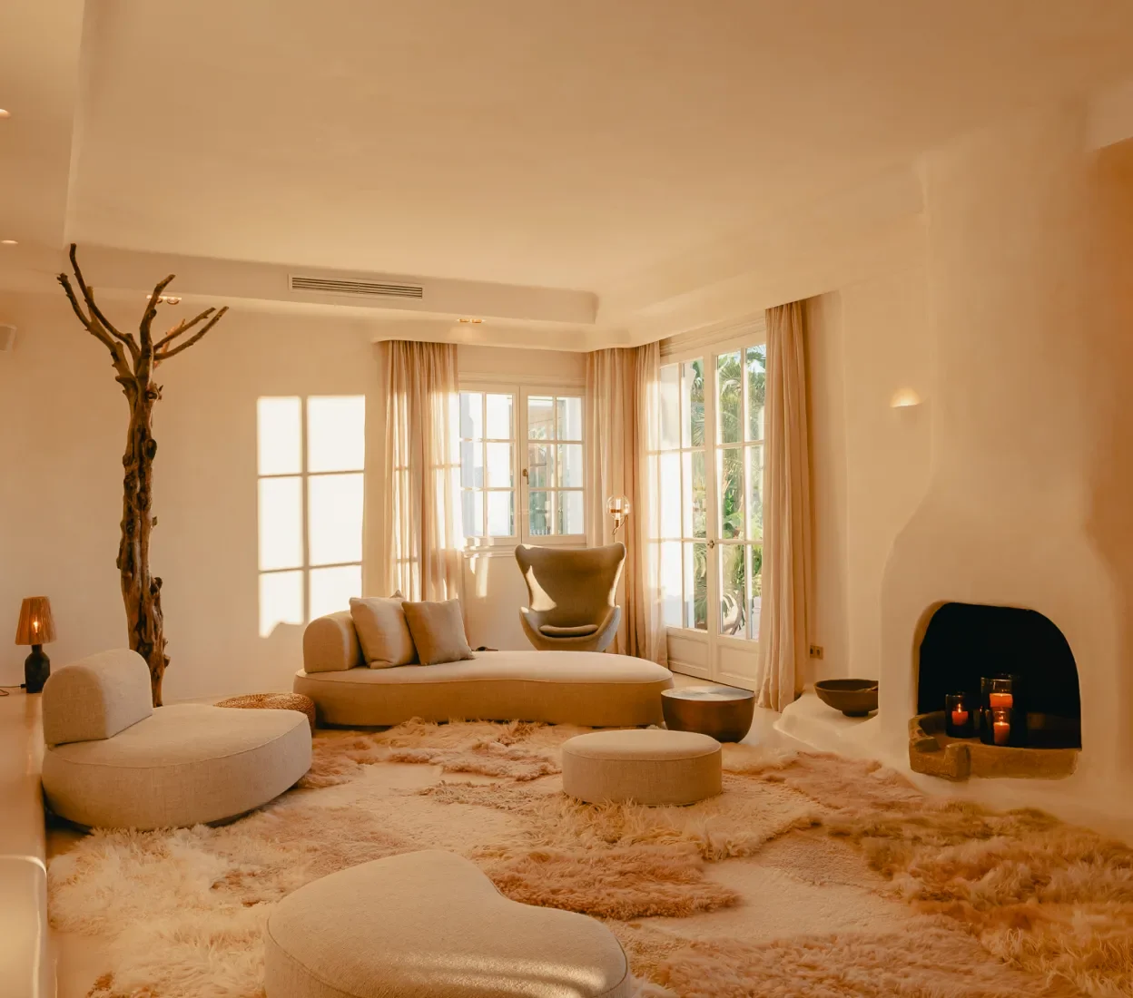 A cozy living room with beige and cream-colored furniture, large windows with curtains, a unique tree-shaped wooden lamp, and a fireplace with candles, bathed in warm sunlight.