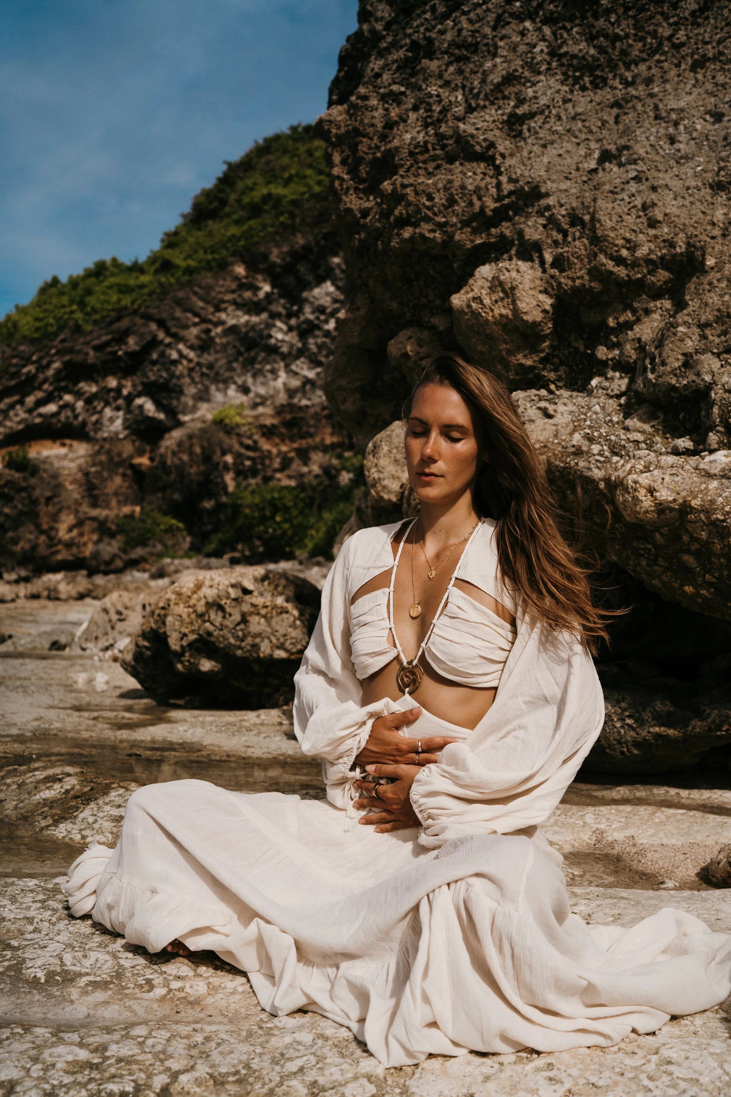 A woman in beige clothing with jewelry, sitting cross-legged on a rocky surface near large rocks and cliffs under a clear blue sky, with her eyes closed and hands resting on her stomach.