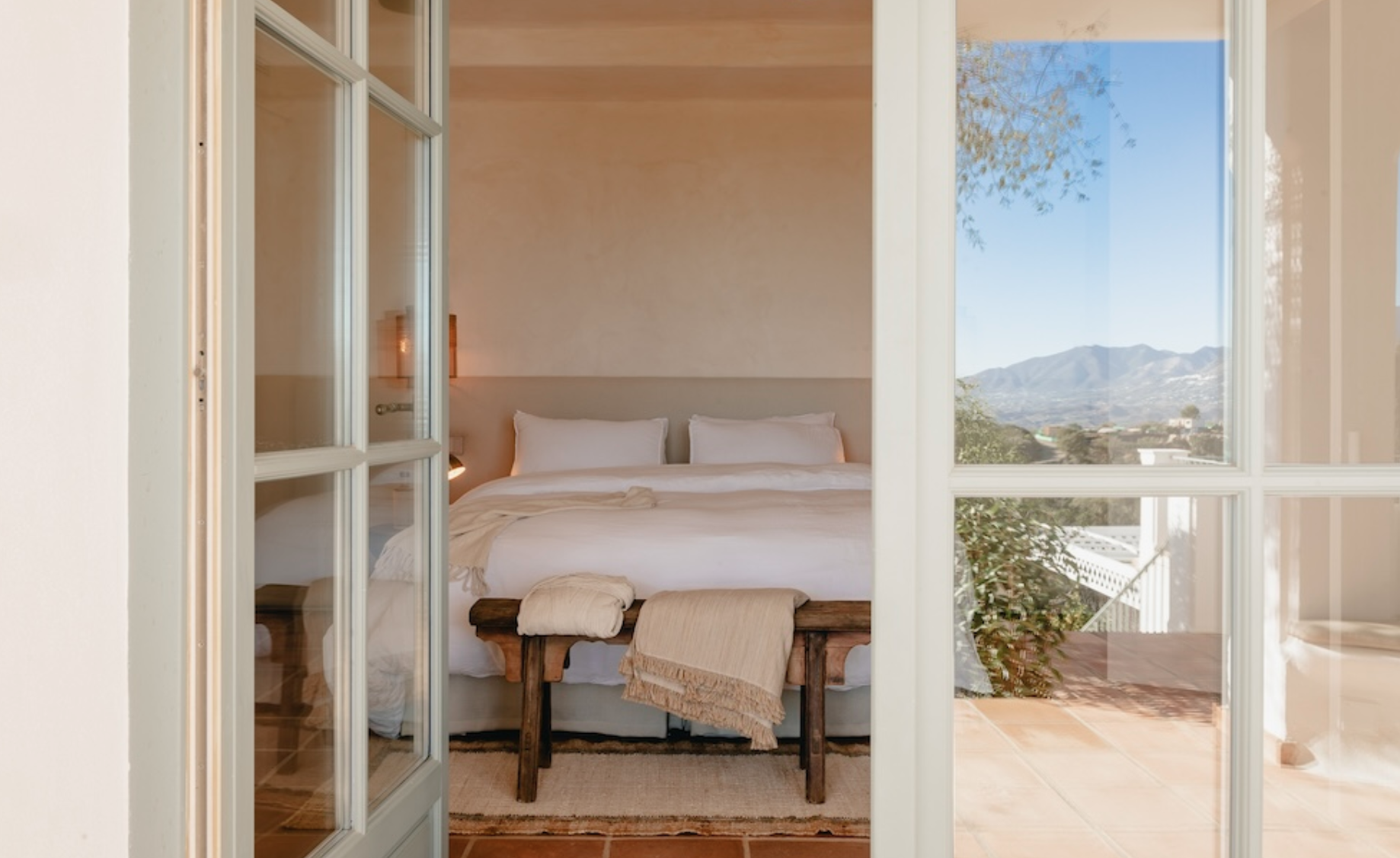 Bedroom with a white bed and natural light, view of mountains and trees through large window.