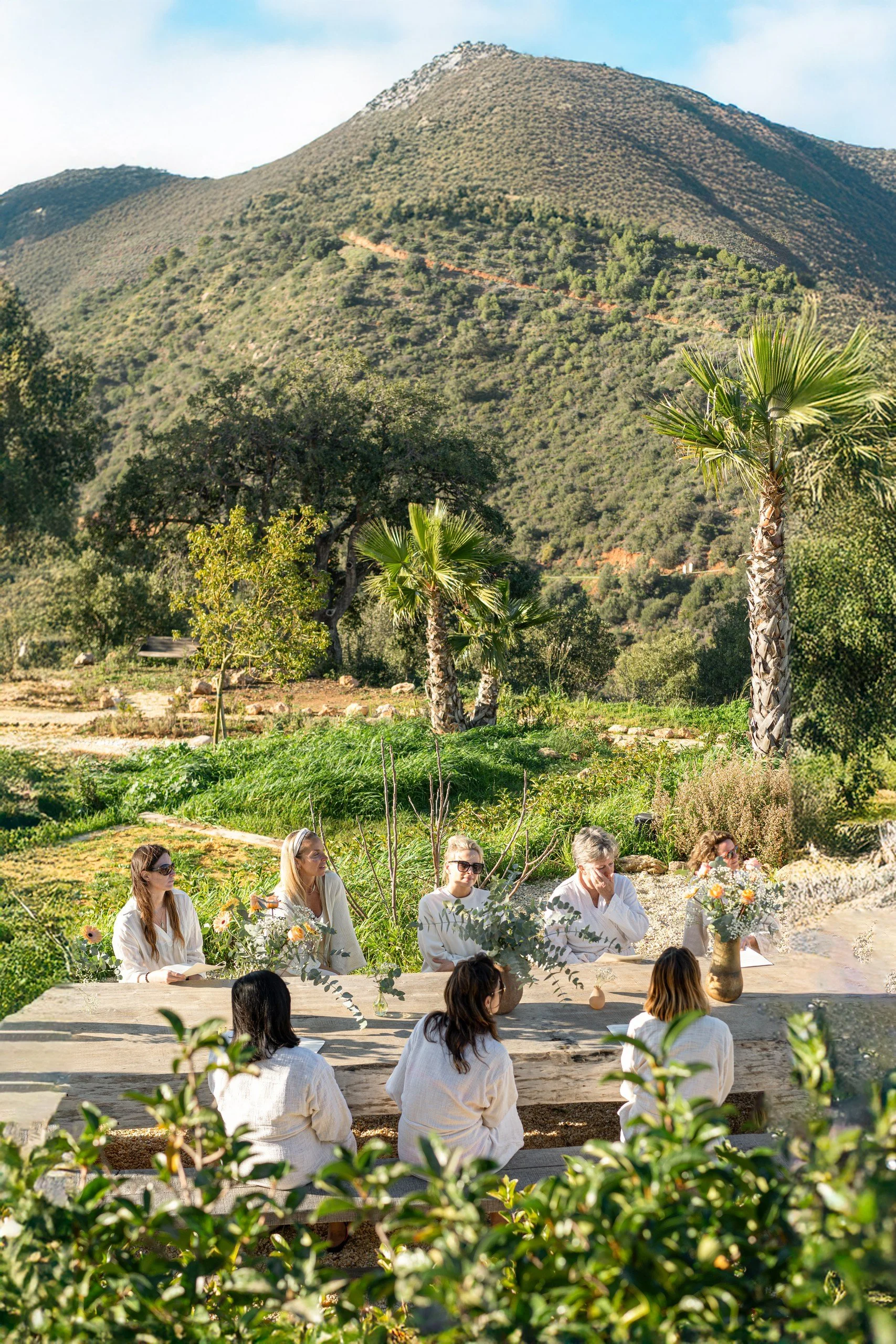 Women seated outdoors at a table with floral arrangements, participating in an event in a scenic mountain setting with trees and hills in the background.