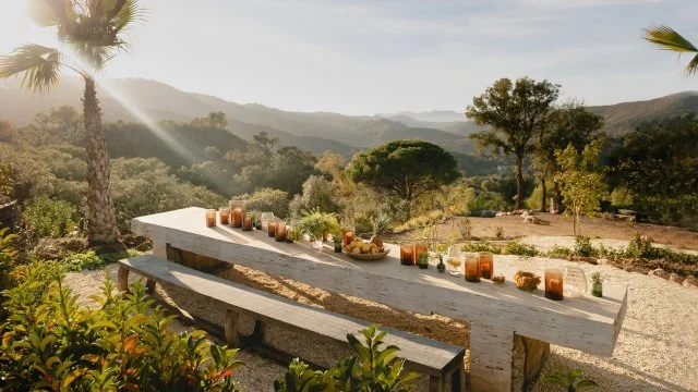 A rustic outdoor dining table with food and drinks, set in a scenic mountain landscape with trees and hills in the background during sunset.