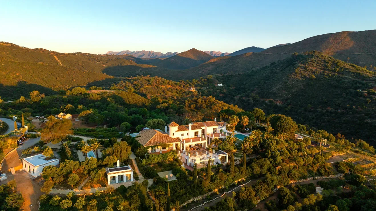 Aerial view of a large house surrounded by trees and mountains during sunset.