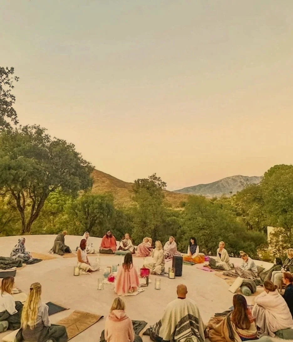 A group of people sitting in a circle outdoors on a yoga deck, surrounded by trees and mountains, participating in a meditation or spiritual session during dusk.