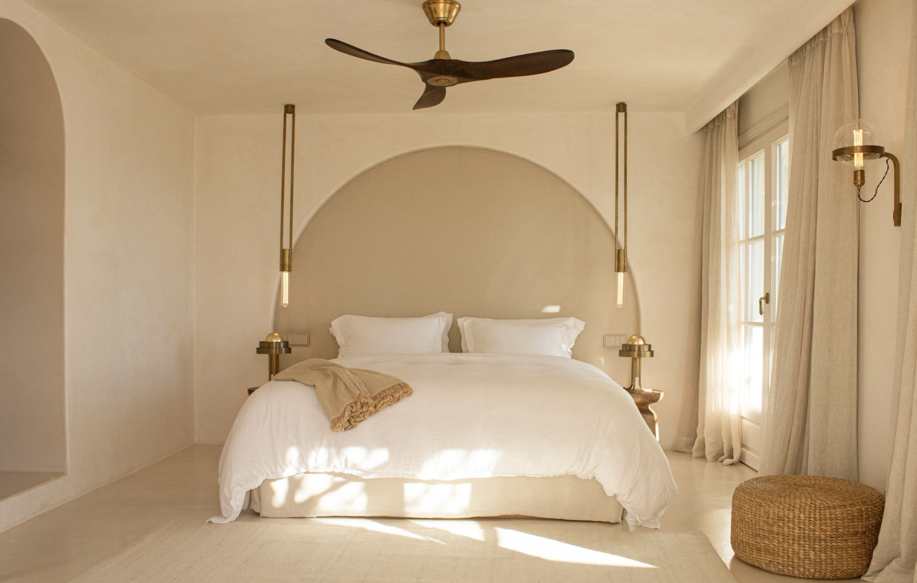 A cozy bedroom with beige walls and natural light coming through a window with light curtains. The bed has white linens and two white pillows, with a beige throw blanket at the foot. Two modern pendant lights hang on either side of the bed, and there are small bedside tables. A wicker ottoman is visible on the right side of the room.