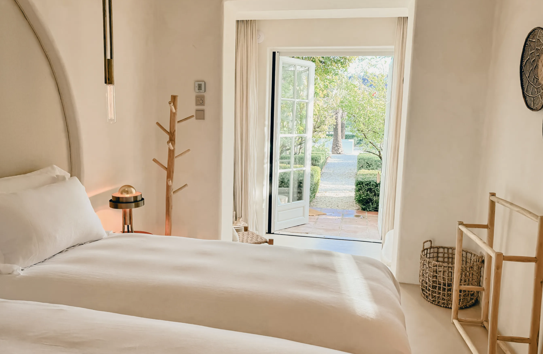 A bedroom with a white bed, a small lamp on a nightstand, a branch-shaped coat rack, and an open door leading to a garden with trees and a pathway.