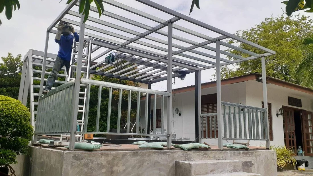 A person wearing a blue shirt and gloves is working on the roof frame of an outdoor porch, which has a metal railing and steps leading up to it. The porch is being built or renovated, with tools and materials around.