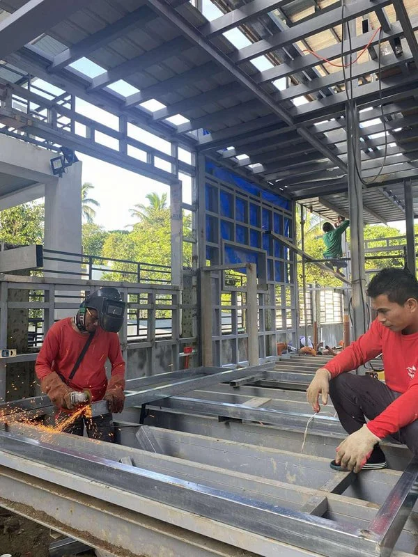 Construction workers assembling metal framework at a building site, with one worker welding and sparks flying, and others working with tools on the structure.