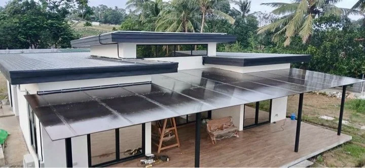 A modern house with solar panels installed on the roof and a balcony with a wooden floor, surrounded by trees and greenery.