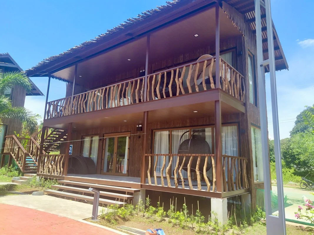 A two-story wooden house with a balcony, stairs leading to the entrance, and wooden railings, surrounded by greenery and clear blue sky.