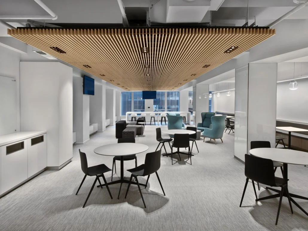 Modern lounge area with round tables, black chairs, blue armchairs, and a wooden ceiling installation in an office space.