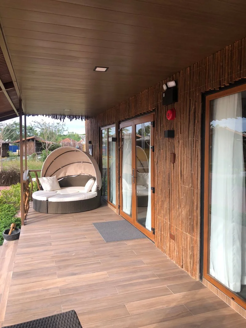 A wooden patio with sliding glass doors, a round daybed with canopy and pillows, a black speaker, and a fire alarm, overlooking a garden with trees and houses in the background.