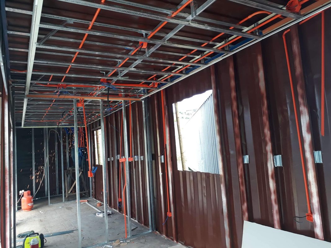 Interior view of a building under construction, showing metal framing, orange electrical conduits, and partially installed wall panels.