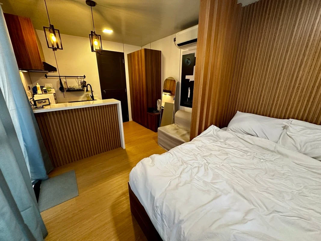 Hotel studio apartment with a bed, a sitting chair, a small kitchen area, and wooden accents.