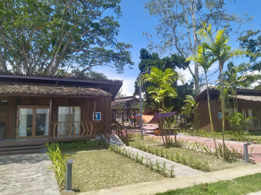 Two wooden cottages numbered 1 and 2 in a tropical garden with palm trees, manicured grass, walking paths, and outdoor seating under shade umbrellas, under a blue sky with trees in background.