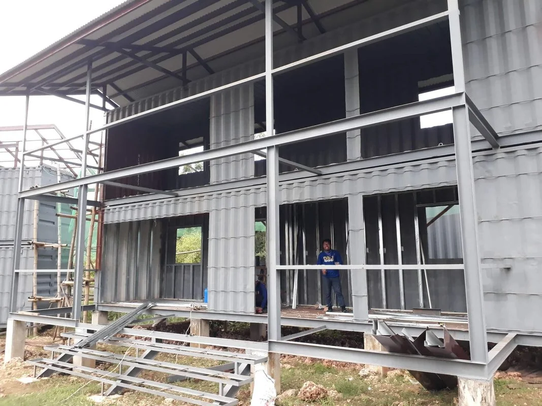 Construction site of a modern, two-story building made from metal sheets with scaffolding and stairs, workers are inspecting the structure.