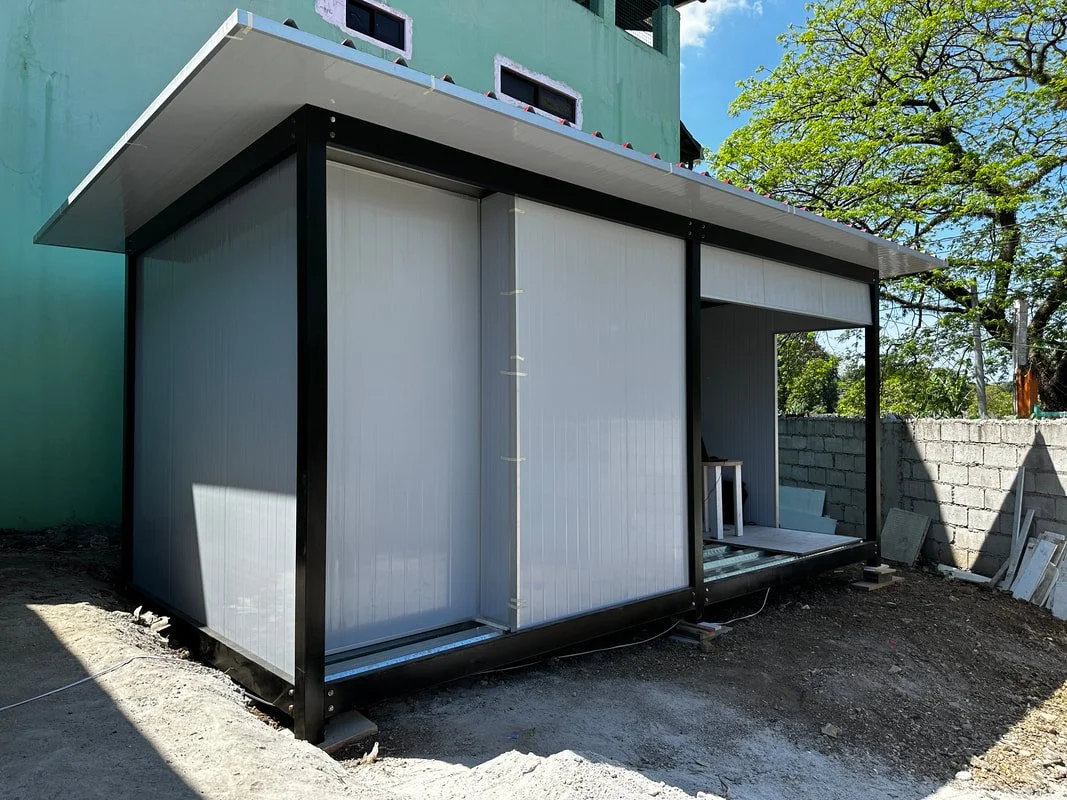 A small, modern, partially constructed building with metal siding, black framing, and a flat roof, situated outdoors next to a tree and a brick wall.