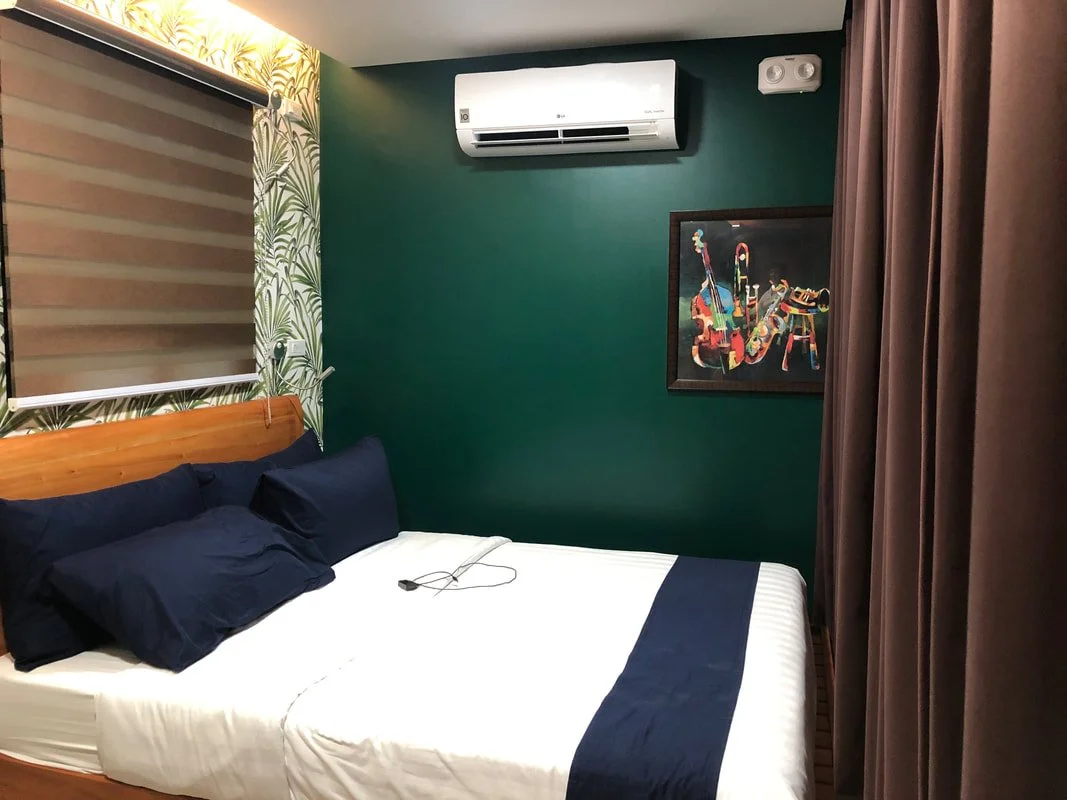 Hotel room with a bed, navy blue pillows, a dark green accent wall, a framed colorful artwork, an air conditioning unit, a window with blinds, and brown curtains.