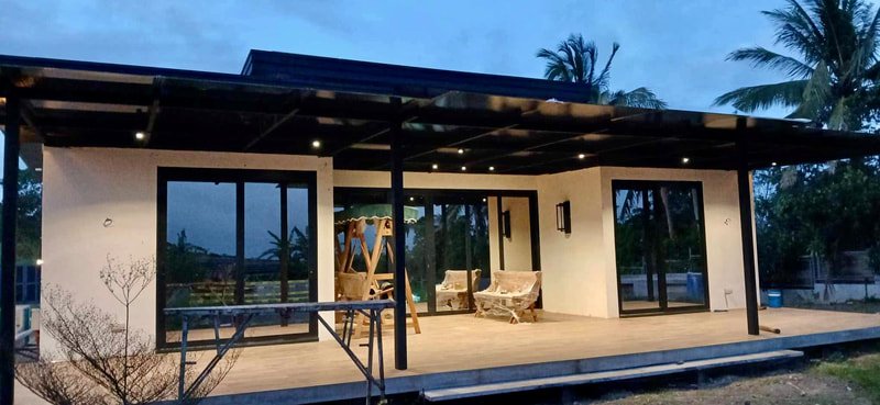 Modern house with a large elevated patio, sliding glass doors, outdoor seating, and a swing, surrounded by palm trees at dusk.