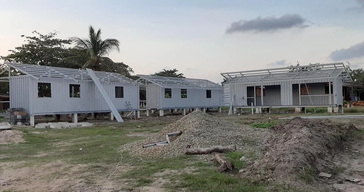 Under construction houses made of shipping containers with metal roofs, surrounded by dirt and construction materials.
