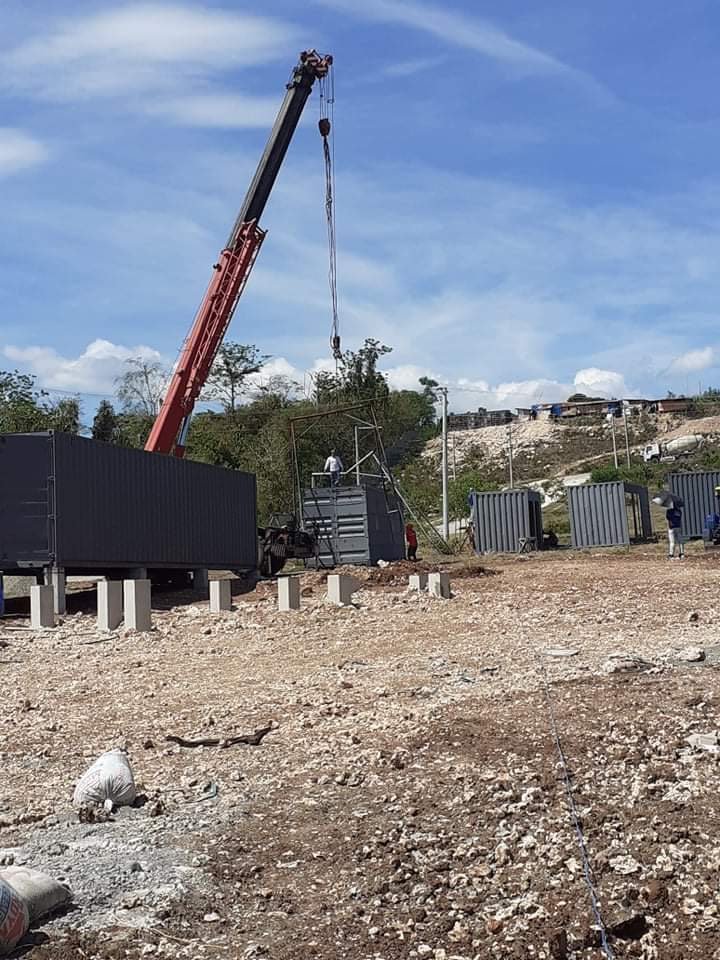 Construction site with a large crane lifting a metal frame, several workers are present, and shipping containers are positioned on uneven, rocky ground.