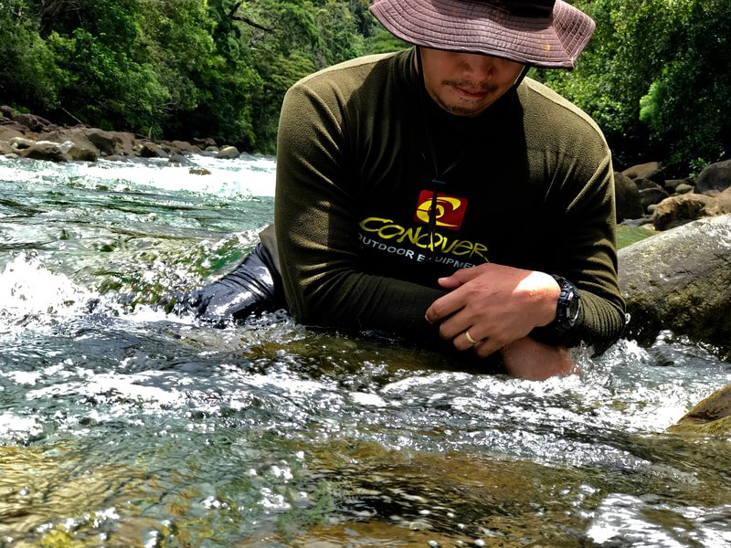 A man wearing a wide-brimmed hat, a dark green long-sleeve shirt, and a wristwatch is kneeling in a river surrounded by rocks and lush green trees, with sunlight filtering through the foliage.