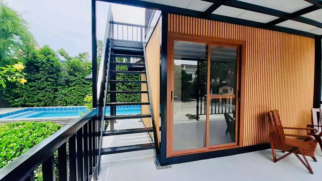 Outdoor patio with a wooden house, sliding glass door, black metal staircase leading to a rooftop deck, pool, and seating area with wooden chairs.