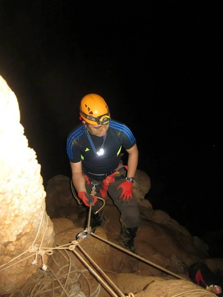 A person wearing a helmet, headlamp, red gloves, and dark clothing is climbing a rocky surface at night with safety ropes and gear.