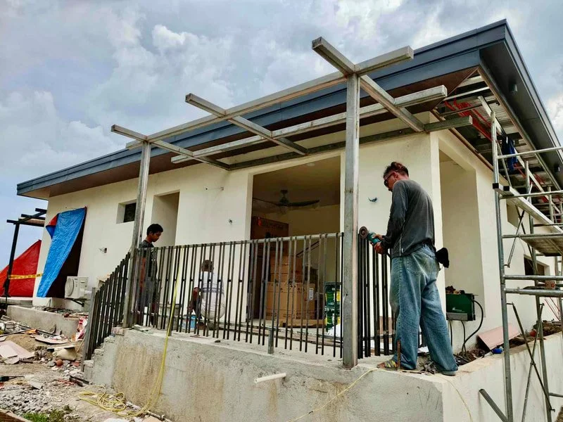 Construction workers building a house, with one using a power tool on a balcony railing, and scaffolding on the side of the house.