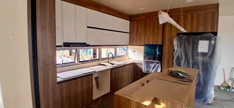 Modern kitchen under construction with wooden cabinets, a long window with multiple small windows, a kitchen island, and appliances covered in plastic.