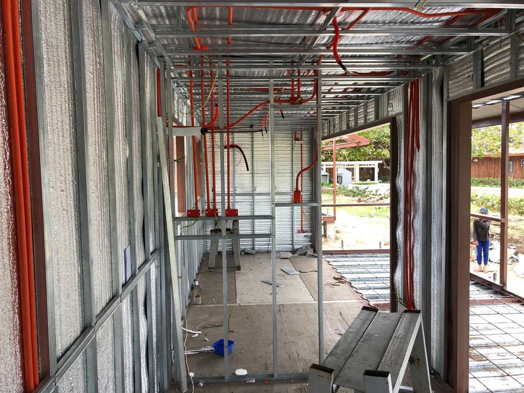 Interior of a building under construction, with exposed metal framing, electrical wiring, and partially installed flooring. Outside, a person is working near the perimeter.
