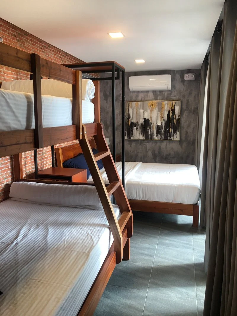 Bedroom with a wooden bunk bed and a single bed, modern decor, artwork on gray wall, air conditioner, brick wall, tile flooring, and curtains.