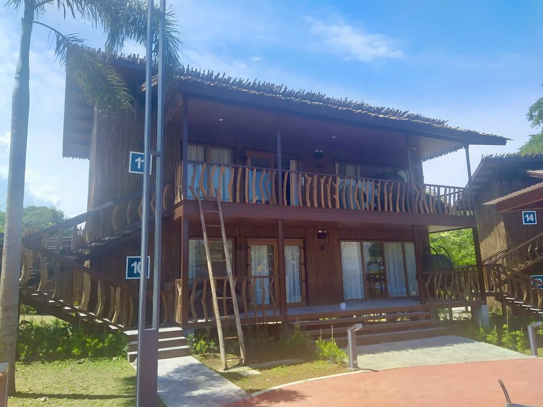 A wooden two-story building with a thatched roof, surrounded by palm trees and greenery, under a partly cloudy sky.