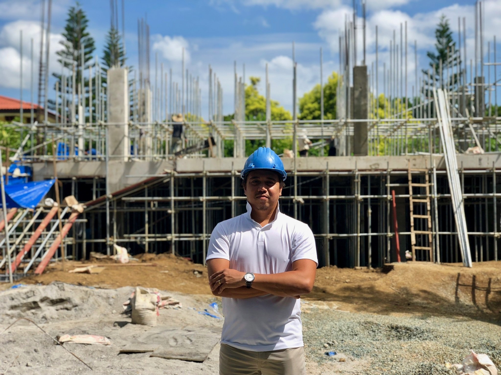 A man wearing a blue safety helmet and white polo shirt stands with arms crossed at a construction site, with a partially built structure in the background and scaffolding around it.