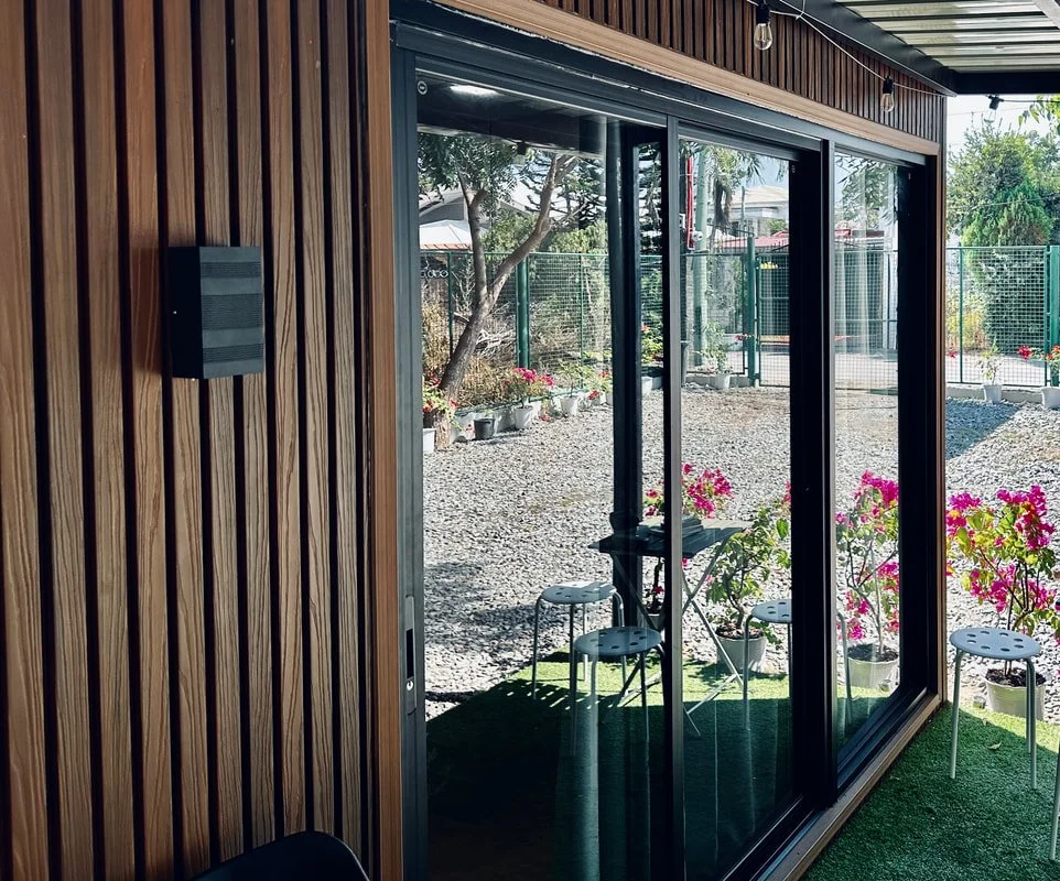View of an outdoor patio with pink flowers, patio furniture, and a gravel ground, seen through large glass sliding doors from inside a building with wooden walls.